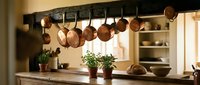 Rustic kitchen interior with copper pots hanging from rack above wooden counter. Warm lighting creates cozy cooking space with herbs and traditional cookware.