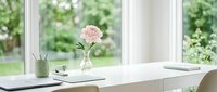 Clean minimalist workspace with pink peony flower in glass vase on white desk by window overlooking green garden for home office interior design.