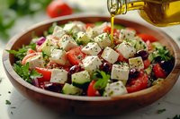 Fresh Greek salad with feta cheese, olives, tomatoes and cucumber being drizzled with golden olive oil in wooden bowl for healthy Mediterranean cuisine.