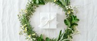 White gift box with ribbon surrounded by fresh green herb wreath on rustic wooden background for natural gifting and celebration occasions.