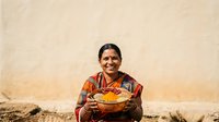 Woman of South Asian descent, middle-aged, wearing traditional colorful embroidered clothing, holding a bowl of turmeric or spices with warm smile against weathered beige wall. Suitable for cultural, culinary, and lifestyle content.