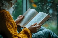 Woman in mustard yellow sweater reading book by window on rainy day, creating cozy atmosphere with raindrops on glass and soft natural lighting.