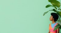 Young child with dark curly hair and medium skin tone wearing colorful clothing, standing against mint green background with tropical plant foliage, looking upward with curiosity and wonder.