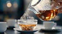 Hot tea pouring from glass teapot into transparent cup with rising steam, dramatic lighting and bokeh background creates cozy atmosphere for tea ceremony.