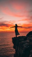 Silhouetted woman standing on rocky cliff with arms outstretched facing vibrant orange and pink sunset over ocean horizon, conveying freedom and contemplation for travel and lifestyle content.
