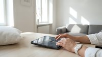 Professional woman using digital tablet in bright modern living room with natural sunlight streaming through windows for remote work and productivity.