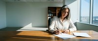 Mature woman with gray hair and glasses wearing white shirt writing at wooden desk in modern office with city view and natural sunlight for business, professional, corporate concepts.