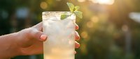Hand holding a refreshing glass of clear lemonade or mojito cocktail with ice and mint leaf garnish outdoors in summer sunlight.
