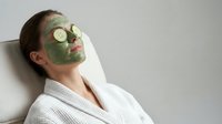 Young Caucasian woman relaxing with green clay facial mask and cucumber slices on eyes, wearing white robe, spa skincare wellness concept.