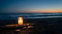 Glowing candle in glass jar on driftwood at ocean beach during sunset twilight with waves and pebbles creating peaceful romantic atmosphere for meditation.