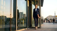Professional blonde businesswoman in navy suit walking through modern glass office building entrance during golden hour sunset with urban cityscape background.