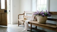 Rustic wooden bench in a bright entryway with a woven basket of purple lilac flowers, straw hat, and soft linen scarf in natural light.