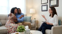 African American couple embracing on couch during therapy session with female counselor in modern office setting for relationship support.