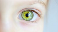 Extreme close-up of bright green human eye with detailed iris pattern and clear pupil, showing delicate eyelashes and pale skin around eye area.