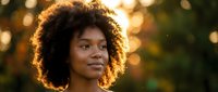Young African American woman with natural curly afro hair smiling outdoors with warm golden hour lighting and bokeh background for lifestyle content.