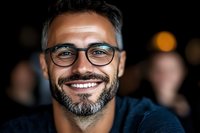 Confident middle-aged man with salt and pepper beard wearing glasses smiles warmly at camera. Dark background with soft bokeh creates professional headshot atmosphere.