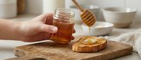 Young woman's hand holding jar of golden honey above slice of bread on wooden board with honey dipper, ceramic bowls and neutral kitchen background, suitable for food, wellness and cooking content.