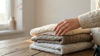 Woman in cream sweater placing hand on stack of folded neutral knit sweaters on wooden table near window in cozy home interior for seasonal wardrobe organization.