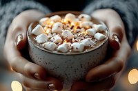 Cozy ceramic mug filled with hot chocolate topped with mini marshmallows and cocoa powder, held in warm hands against blurred holiday lights background.