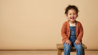 Young child with curly brown hair and mixed ethnicity, wearing rust-colored cardigan and denim overalls, sitting on wooden stool against neutral beige background, smiling at camera for portrait or advertising use.