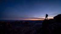 Silhouette of hiker standing on rocky cliff edge during dramatic sunset with purple and orange sky over desert canyon landscape for adventure travel.