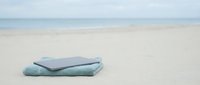 Folded light blue towels resting on sandy beach with calm ocean and horizon in background, suitable for travel, vacation, wellness, and resort marketing materials.