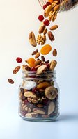Mixed nuts and dried fruits pouring from glass jar onto white surface. Almonds, walnuts, pecans, cashews, raisins, cranberries and apricots for healthy snacking and nutrition.