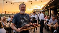 Cheerful bearded chef presenting grilled meat platter at busy outdoor restaurant during golden hour sunset with string lights and dining guests.