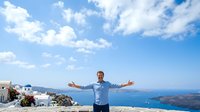 Man with arms outstretched standing on cliff overlooking blue sea and white-walled buildings, expressing freedom and joy during travel vacation.