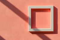 White square frame mounted on coral pink wall casting dramatic diagonal shadow in sunlight, creating minimalist geometric composition with depth effect.