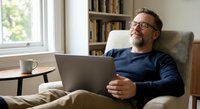 Middle-aged Caucasian man with beard and glasses relaxing in armchair with laptop at home. Remote work, freelancing, comfortable lifestyle concept.