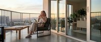 Relaxed businessman in casual attire sitting on modern balcony chair during golden hour sunset, enjoying peaceful moment with city skyline view from luxury apartment.