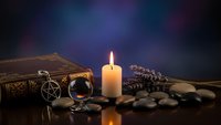 Mystical fortune telling setup with candle, crystal ball, ancient book, lavender and stones on dark surface with purple blue gradient background.