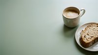 Warm cup of coffee with latte foam beside fresh whole grain bread slice on plate, minimalist breakfast setup on soft green surface for morning beverage and food styling.