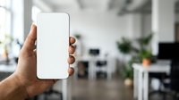 Man holding smartphone with blank white screen in modern office workspace with plants and natural light.