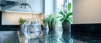 Fresh water pouring from a pitcher into a glass on a dark granite kitchen counter with green plants in the background.