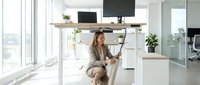 Young woman with light brown hair and fair complexion, wearing casual beige clothing, sitting at a standing desk in a bright, modern office space with large windows, working on a laptop in a contemporary workspace.
