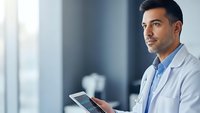 Professional male doctor in white coat holding digital tablet in modern medical office environment for healthcare technology applications.
