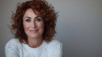Confident middle-aged woman with curly red hair wearing white knit sweater against neutral gray background, smiling warmly at camera.