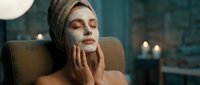 Young Caucasian woman with towel on head applying white clay face mask, relaxing in spa atmosphere with candles, enjoying skincare beauty routine.