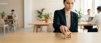 Young professional woman of South Asian descent in dark blazer placing wooden house model on wooden table in modern office environment with colleagues working in background.