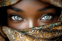 Striking blue eyes peek through traditional patterned fabric wrap, creating dramatic contrast with dark skin. Artistic portrait highlighting cultural beauty and mystery.