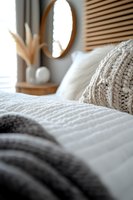 Cozy bedroom interior with white bedding, knitted throw pillows, and round mirror on wall. Soft morning light creates peaceful atmosphere for lifestyle decor.