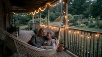Mature couple relaxing in hammock on wooden deck at dusk, surrounded by warm string lights and lush garden views, perfect for lifestyle and home decor content.