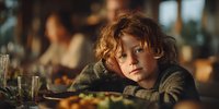 Pensive young boy with curly hair resting head on table during family dinner, warm atmospheric lighting creates emotional mood for family dynamics storytelling.