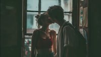 Young couple sharing intimate moment by window in cozy apartment with warm lighting and urban view for romantic lifestyle content.