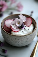 Ceramic bowl with creamy yogurt garnished with sliced radishes, delicate edible flowers, and purple herbs creating an artistic food composition on marble surface.