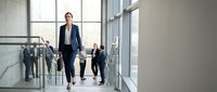 Confident businesswoman walking in modern office corridor with team members in background, corporate leadership and professional workplace environment.