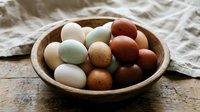 Fresh farm eggs in various natural colors displayed in rustic wooden bowl on weathered wood surface with linen cloth for organic cooking and healthy eating.