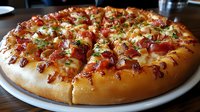 Fresh baked pizza with crispy bacon bits, melted mozzarella cheese and herbs on thick golden crust, served on white plate in restaurant setting.